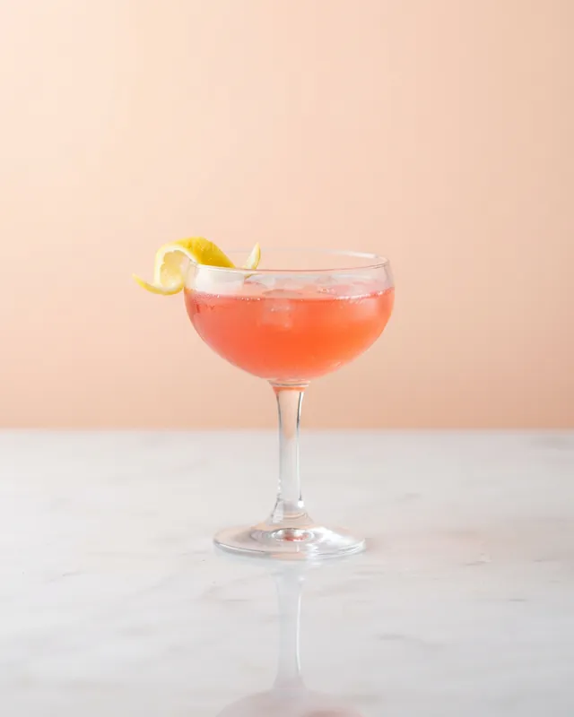 A coral-pink cocktail with a lemon twist in a coupe glass against a pastel peach backdrop