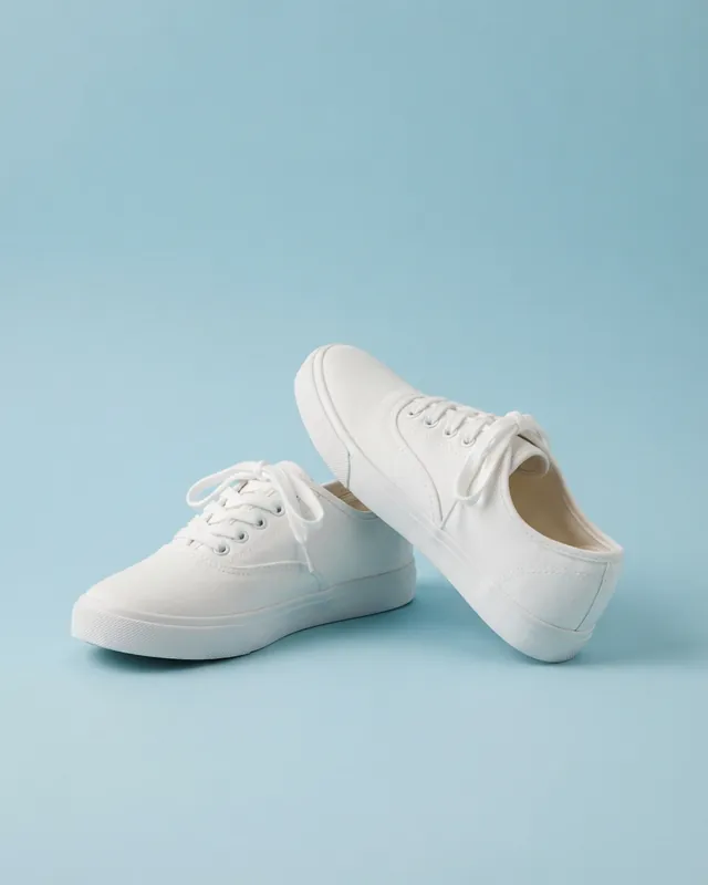 A pair of plain white canvas sneakers on a pastel blue backdrop, one leaning against the other