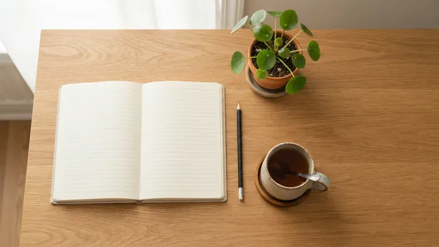 Top-down view of an oak desk with an open lined notebook, a pencil, a cup of tea and a small potted plant