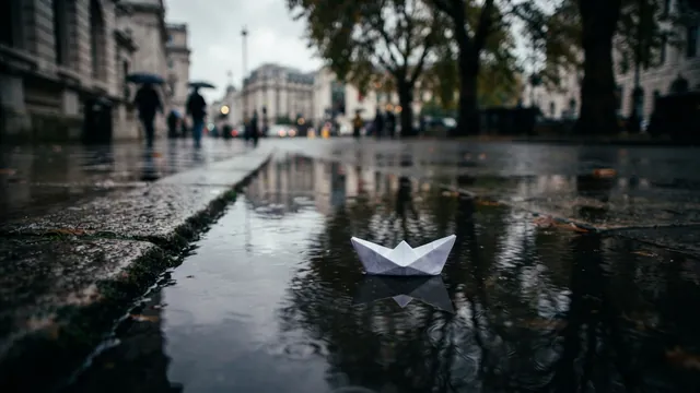 A white paper boat floating in a rain puddle on a city sidewalk, with blurred people under umbrellas behind