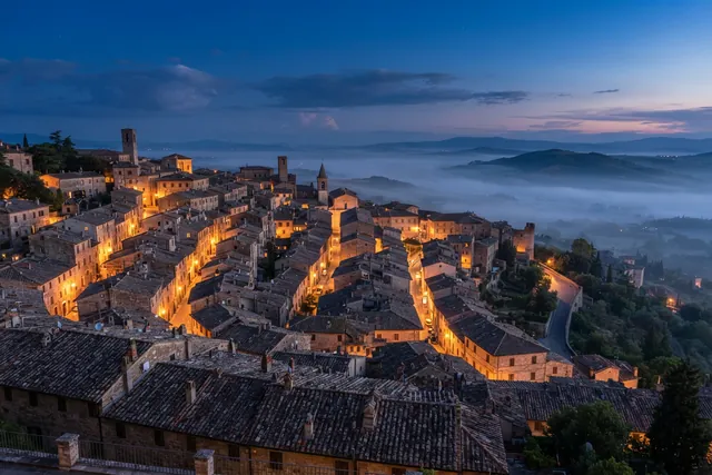 Rooftops of an old hillside town at blue hour, with lit streets and mist in the valley beyond